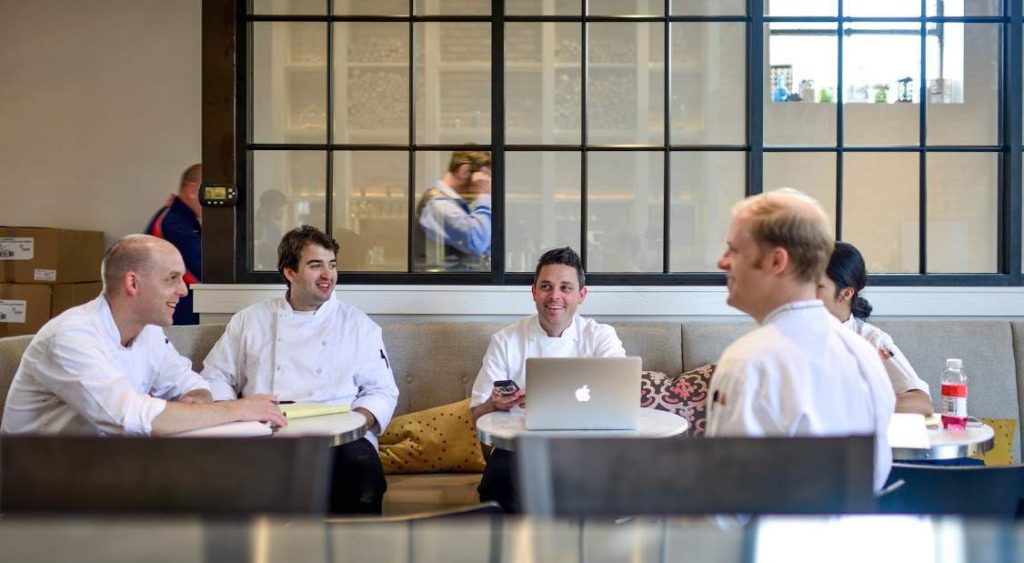 The image shows three chefs sitting at a restaurant table while working on their laptops.