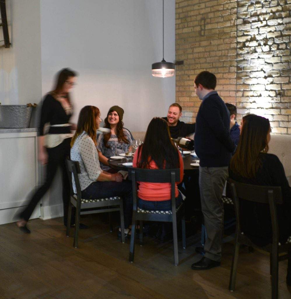 The image shows a restaurant setting. A group of friends are sat at a table and two servers are brining them dishes and pouring wine.