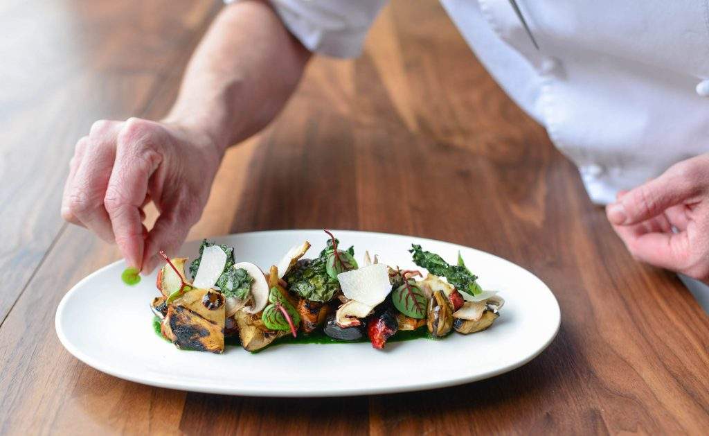 The image shows a chef putting in the final touch before serving a dish. On the plate, there are several veggies and leafy greens.