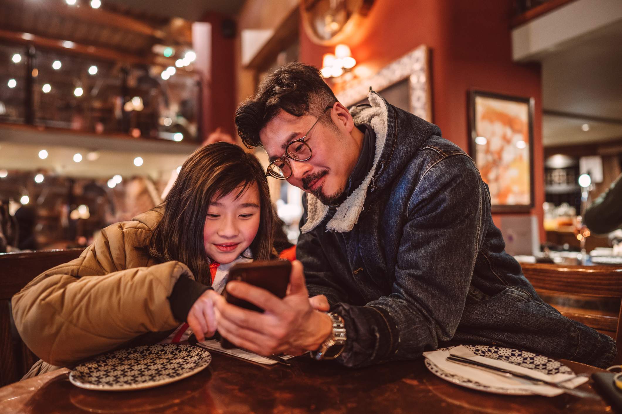 Image depicts a child and their parent using a phone together while sitting at a table in a restaurant. 