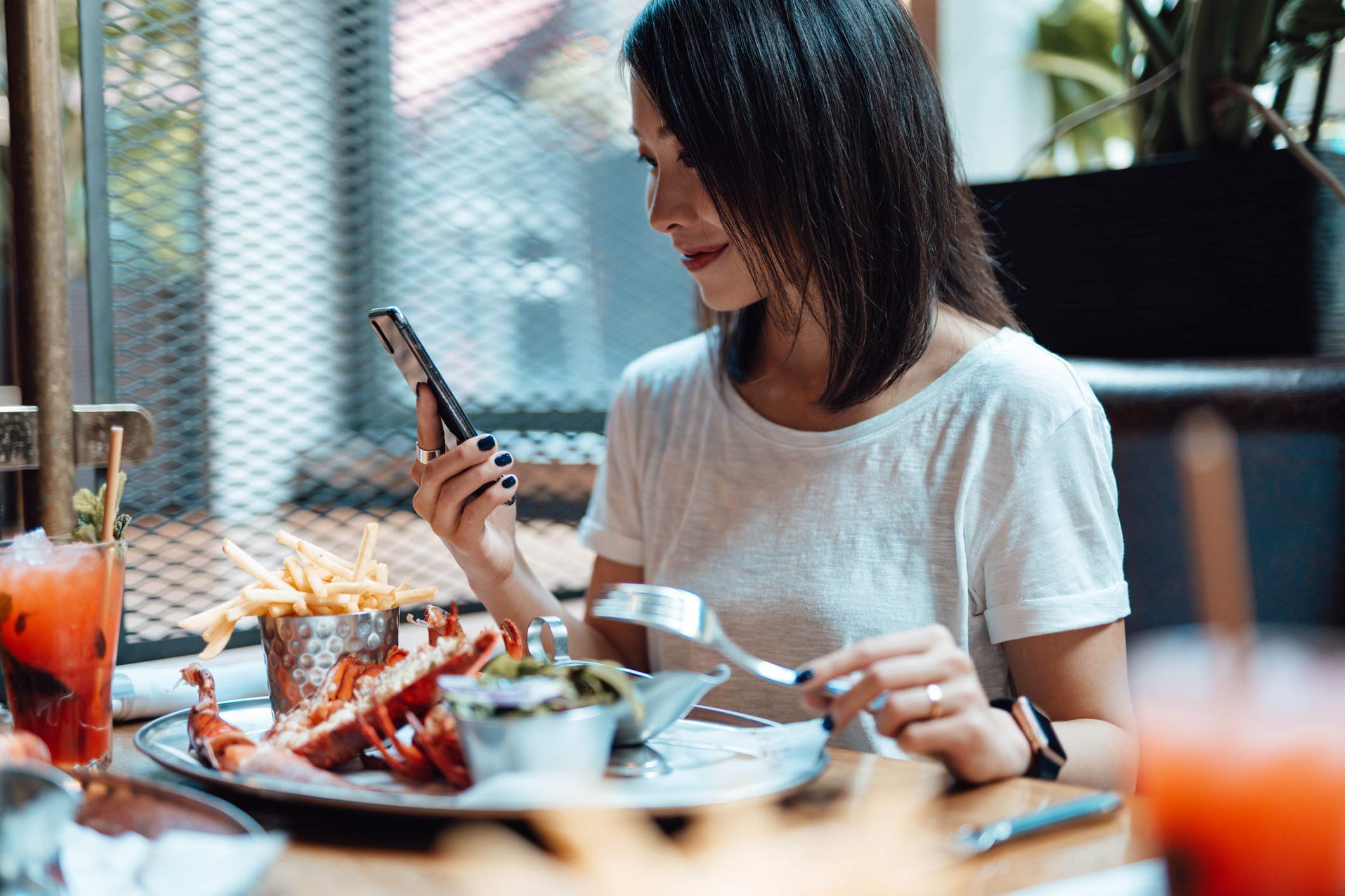 Image depicts a person smiling at their phone while seated at a restaurant. There is a colorful drink in front of them and a plate of seafood and fries. 