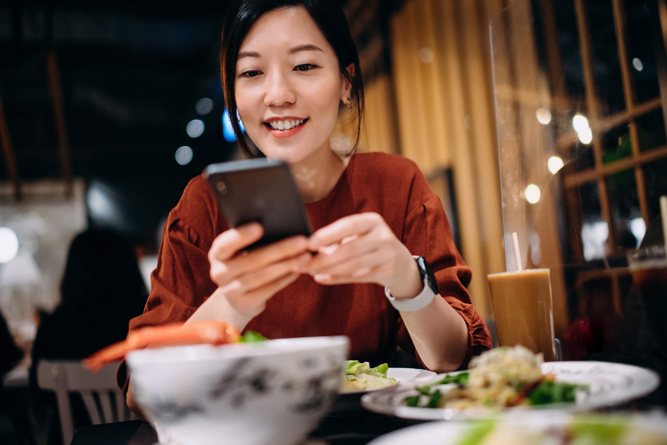 l'image montre une personne dans un restaurant utilisant son téléphone. Elle porte une chemise rouge et une smartwatch et elle sourit en regardant son téléphone.