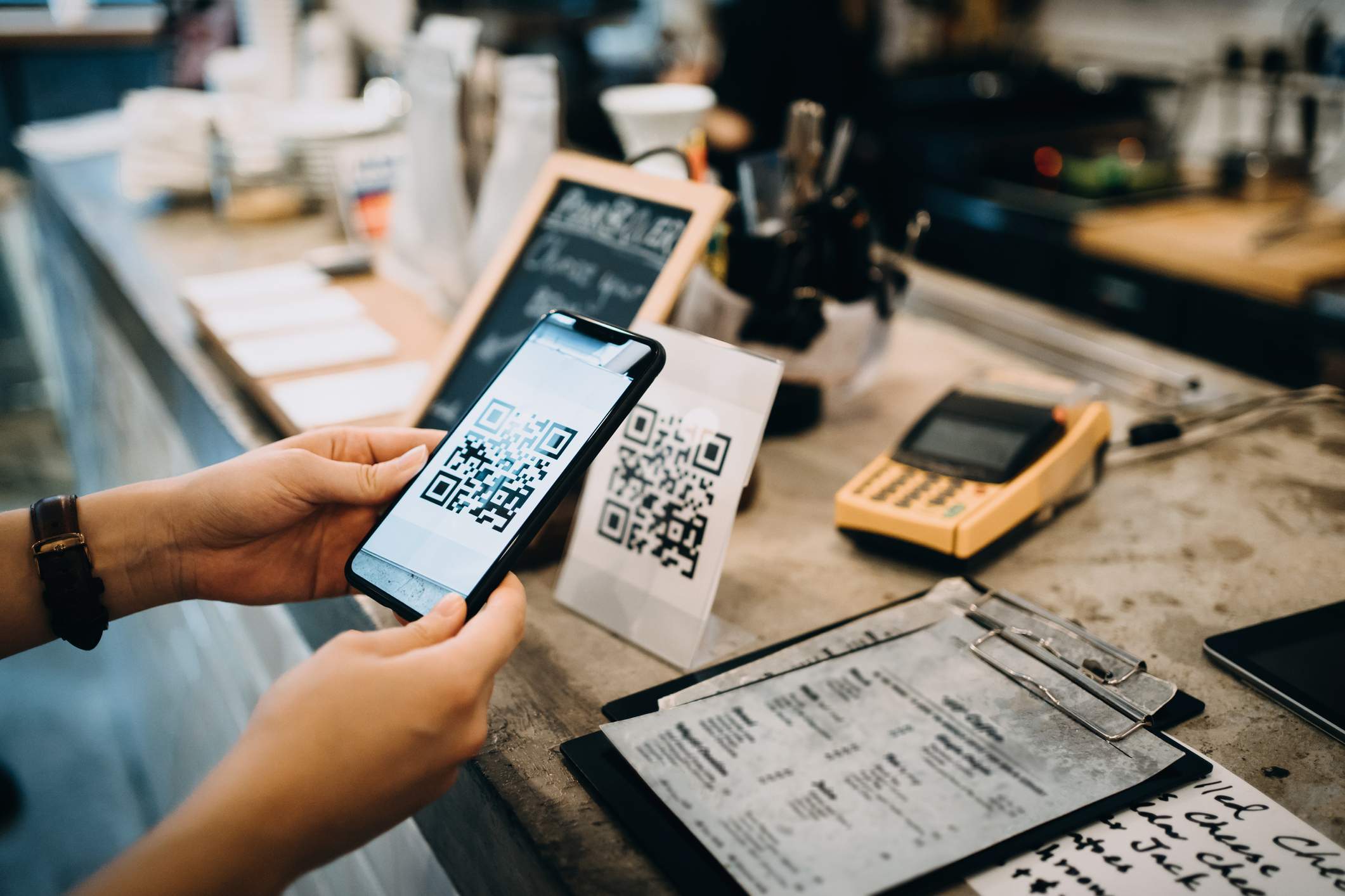 L'image représente une personne tenant un téléphone devant le comptoir d'un restaurant et scannant un code QR.